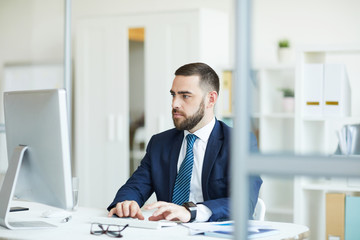 Serious thoughtful young bearded businessman in dark blue suit sitting at table and typing on...