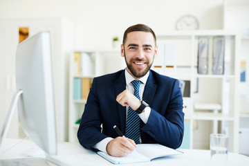 Portrait of happy excited young bearded CEO in formal suit sitting at table with computer and writing down goals in diary