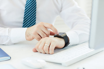 Close-up of unrecognizable businessman in white shirt sitting at desk with computer and reading message on smartwatch