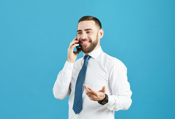 Smiling optimistic young bearded manager in white shirt and stripped tie standing against blue wall and talking to customer on cellphone