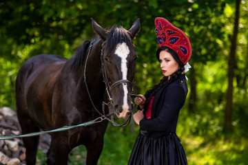 Fototapeta premium Fashion Model in Russian Kokoshnik Holding Thoroughbred Horse Against Nature Background.