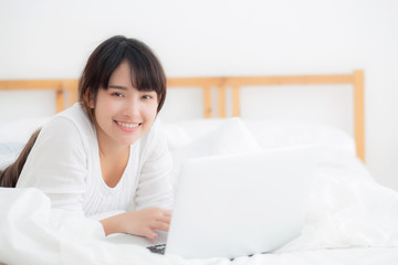 Beautiful portrait asian young woman smile lying on bed using laptop computer with happy at bedroom for leisure and relax, freelance with girl working notebook, communication concept.