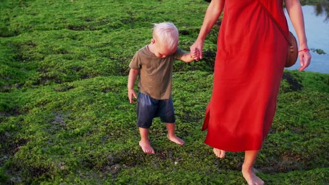 Barefooted mother and son are walking carefully on green algae at low tide. Famous Balangan beach, Bali, Indonesia