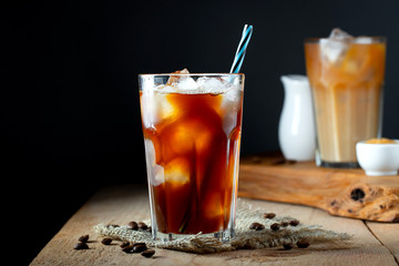 Ice coffee in a tall glass with cream poured over, ice cubes and beans on a old rustic wooden table. Cold summer drink with tubes on a black background with copy space