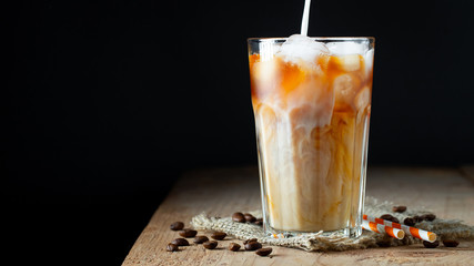 Ice coffee in a tall glass with cream poured over, ice cubes and beans on a old rustic wooden...