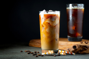 Ice coffee in a tall glass with cream poured over, ice cubes and beans on a old rustic wooden table. Cold summer drink with tubes on a black background with copy space