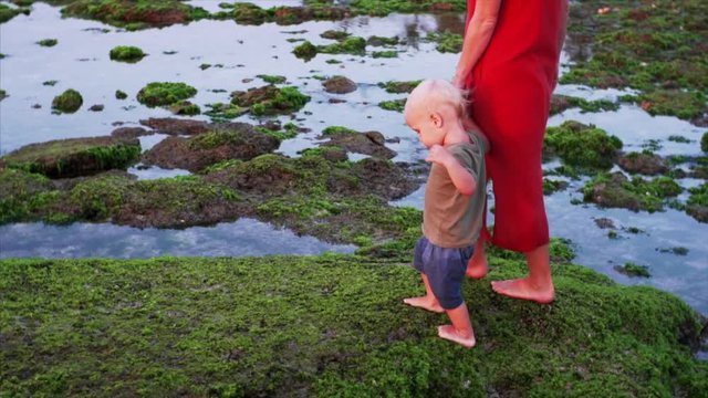 Slow motion of barefooted caucasian family, mother and cute son walking on green algae, exploring shore at low tide. Famous Balangan beach, Bali, Indonesia