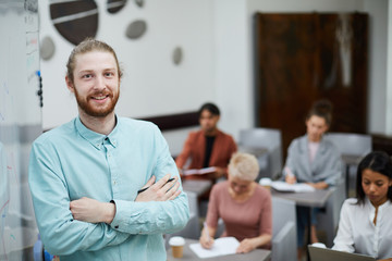Waist up portrait of contemporary male teacher smiling at camera while standing by whiteboard with...
