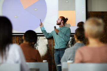 Portrait of contemporary bearded man giving presentation standing by projector screen in lecture...