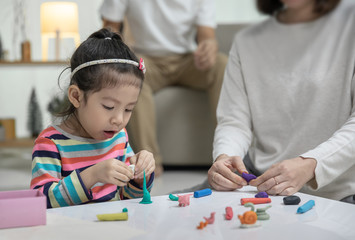 Mom with her daughter playing with a colorful toys modeling clay on the floor, at background daddy is sitting in a sofa in the living room, Happy asian family at home