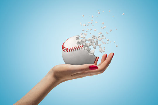 Female hand holding small baseball ball shattering into pieces on blue background