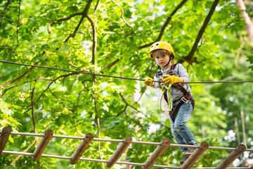 Naklejka na ściany i meble Beautiful little child climbing and having fun in adventure Park. Toddler kindergarten. Children summer activities. Child climbing on high rope park. Small boy enjoy childhood years