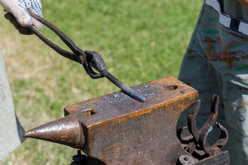 Blacksmith forges metal billet on the anvil against the background of green grass
