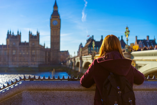 Back view of tourist ginger girl in purple coat with blur Big Ben in the background on beautiful blue sky sunny day at Westminster Bridge, England