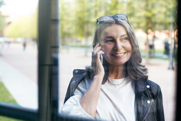 Picture of attractive fashionable middle aged businesswoman in stylish leather jacket smiling broadly while talking using cell phone. View through window glass of mature female using mobile
