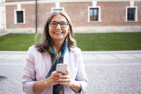 Outdoor picture of good looking stylish European middle aged woman using 4g wireless internet connection on her cell phone texting messages using online messenger, staying always connected