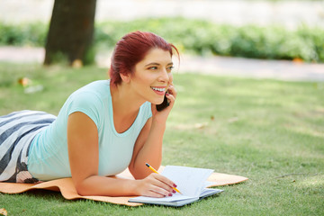 Fototapeta na wymiar Happy attractive young woman sketching in notepad when talking on phone with friend