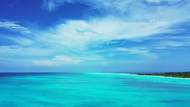 Aerial view of the turquoise blue sea in the Maldives. With an island under a cloudy sky. Pan left.