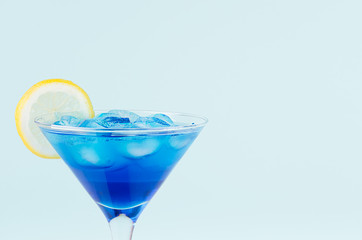 Blue curacao cocktail with ice cubes, lemon slice in luxury martini glass on pastel mint color wall, closeup, edge, details.
