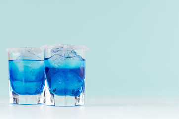 Refreshing alcohol drinks blue Hawaii in two misted elegant shot glass with ice cubes and salt rim in modern pastel mint beach bar interior.