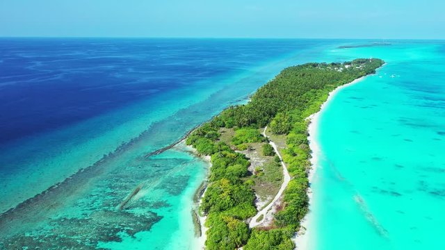 Aerial view, flying over a thin tropical island. A forest on it and a village at the other end. Dolly in. Surrounded by the turquoise sea.