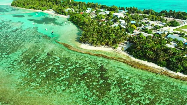 Aerial view flying above tropical island with a village on it.  The turquoise green sea of the Gulf of Thailand in the foreground and reefs in it.. Dolly out