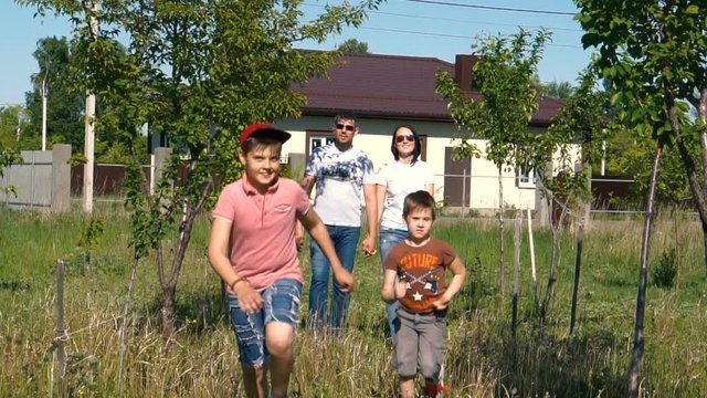 A young family of four. Walk in the garden on a country plot. Good mood. Green meadow and houses in the background. Bright summer day. FullHD