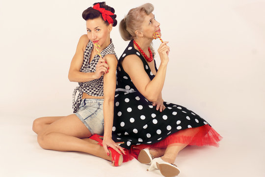 Two pinup girlfriends are sitting with their backs to each other and holding Christmas lollipops on a white background