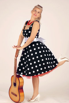 Beautiful blond woman in pinup style, dressed in a polka-dot dress, stands and flirtatiously holds an acoustic guitar in front of her, white background