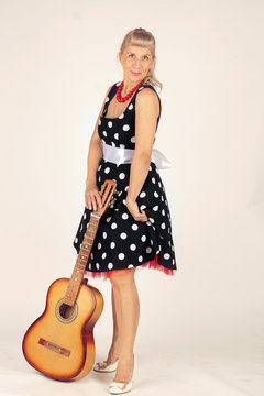 Beautiful blond woman in pinup style, dressed in a polka-dot dress, stands and holds an acoustic guitar in front of her, white background