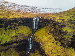 Aerial footage of Fossa waterfall from above during early spring with snow-covered mountain peaks and lush green tones (Faroe Islands, Denmark, Europe)