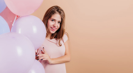 Studio shot of relaxed european girl celebrating her birthday. Indoor portrait of stunning young lady isolated on light background with party balloons.