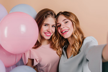 Close-up portrait of excited girl with curly hair making selfie with friend. Indoor photo of charming female models posing with pink balloons.