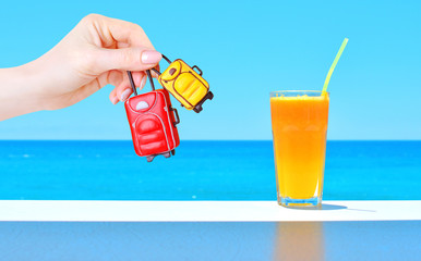 Miniature toy baggage in female hand and orange fresh juice in a glass on the table with blue sea backdrop. Travel concept.