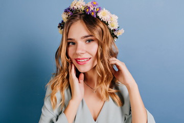 Spectacular lady with pink lips and shiny hair posing on dark background. Adorable girl in flower wreath standing near blue wall.