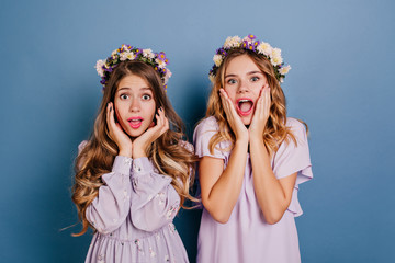 Close-up portrait of two winsome ladies posing on blue background with surprised face expression. Indoor photo of girls with flowers in hair expressing amazement.