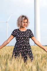Fototapeta na wymiar young tanned curly haired woman in dress is happy to be on the field of ripened wheat, wind turbine on the background