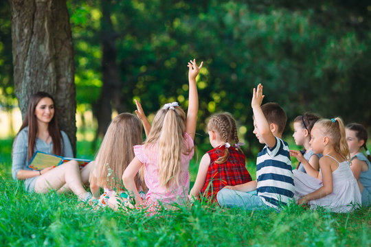 Children and education, young woman at work as educator reading book to boys and girls in park.