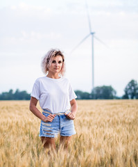 young tanned curly haired woman in white t-short and denim shorts is happy to be on the field of ripened wheat, wind turbine on the background