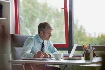 Portrait of a senior business man working in his office, he is taking notes on a pad of paper and using a pen whilst looking at his laptop screen