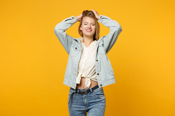 Fototapeta premium Portrait of stunning young woman in denim casual clothes standing, putting hands on head, keeping eyes closed isolated on yellow orange wall background. People lifestyle concept. Mock up copy space.