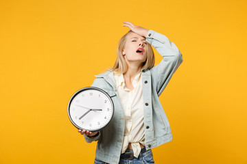 Portrait of exhausted young woman in denim casual clothes putting hand on head, holding round clock...