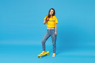 Portrait of cheerful young woman in vivid casual clothes looking camera and standing with yellow skateboard isolated on blue wall background in studio. People lifestyle concept. Mock up copy space.