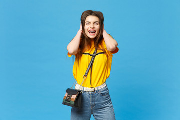 Portrait of laughing young woman in vivid casual clothes with cross body bag putting hands on head...