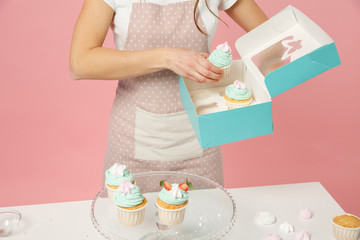 Close up cropped housewife female chef cook confectioner or baker in apron white t-shirt packaging cake cupcake at table isolated on pink pastel background in studio. Mock up copy space food concept.
