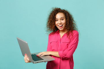 Portrait of excited african girl in pink casual clothes using laptop pc computer isolated on blue...