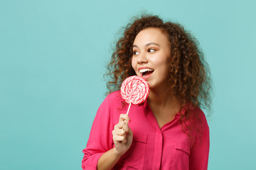 Young african girl in casual clothes holding, eating pink round lollipop, looking aside isolated on blue turquoise background in studio. People sincere emotions, lifestyle concept. Mock up copy space.