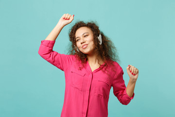 Fototapeta premium Funny pretty african girl in pink casual clothes listening music with headphones and dancing isolated on blue turquoise wall background. People sincere emotions, lifestyle concept. Mock up copy space.