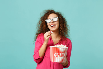 Laughing african girl in 3d imax glasses watching movie film and holding popcorn isolated on blue...