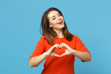 Beautiful brunette young woman wearing red orange dresss howing shape heart sign with hands, isolated over blue wall background, studio portrait. People lifestyle fashion concept. Mock up copy space.